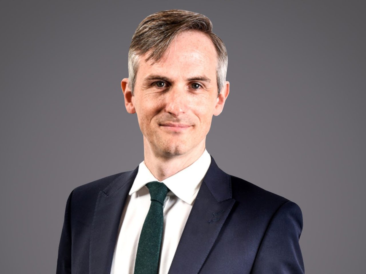 Head-and-shoulders portrait of a person in a navy suit, white shirt and dark green tie, smiling softly against a neutral grey background.