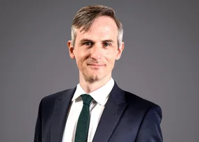 Head-and-shoulders portrait of a person in a navy suit, white shirt and dark green tie, smiling softly against a neutral grey background.