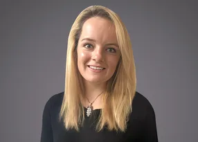Person with long blonde hair, smiling, wearing a black top and a silver owl pendant, against a grey gradient background.