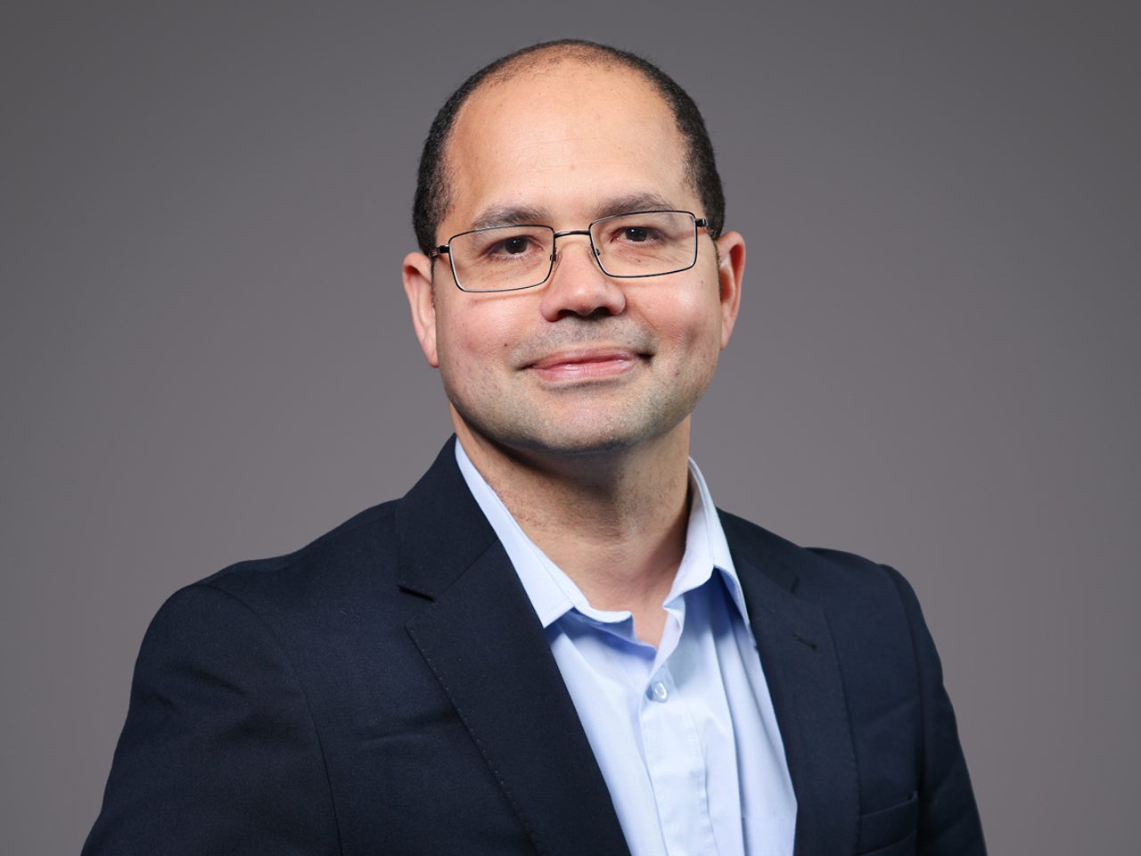 A person with short hair and glasses wearing a dark navy blazer and light blue shirt, smiling softly against a plain gray studio background.