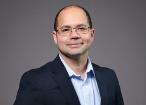 Portrait of a person with short hair and glasses, wearing a navy blazer and light blue shirt, smiling softly against a grey studio backdrop.