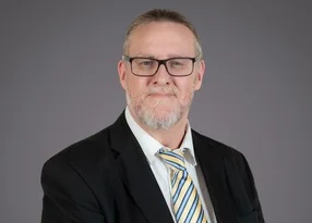 Portrait of a person with grey hair and beard, wearing glasses, a white shirt, striped yellow and blue tie, and a black blazer against a grey backdrop