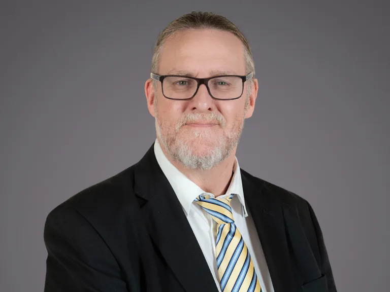 Portrait of a person with grey hair and beard, wearing glasses, a white shirt, striped yellow and blue tie, and a black blazer against a grey backdrop