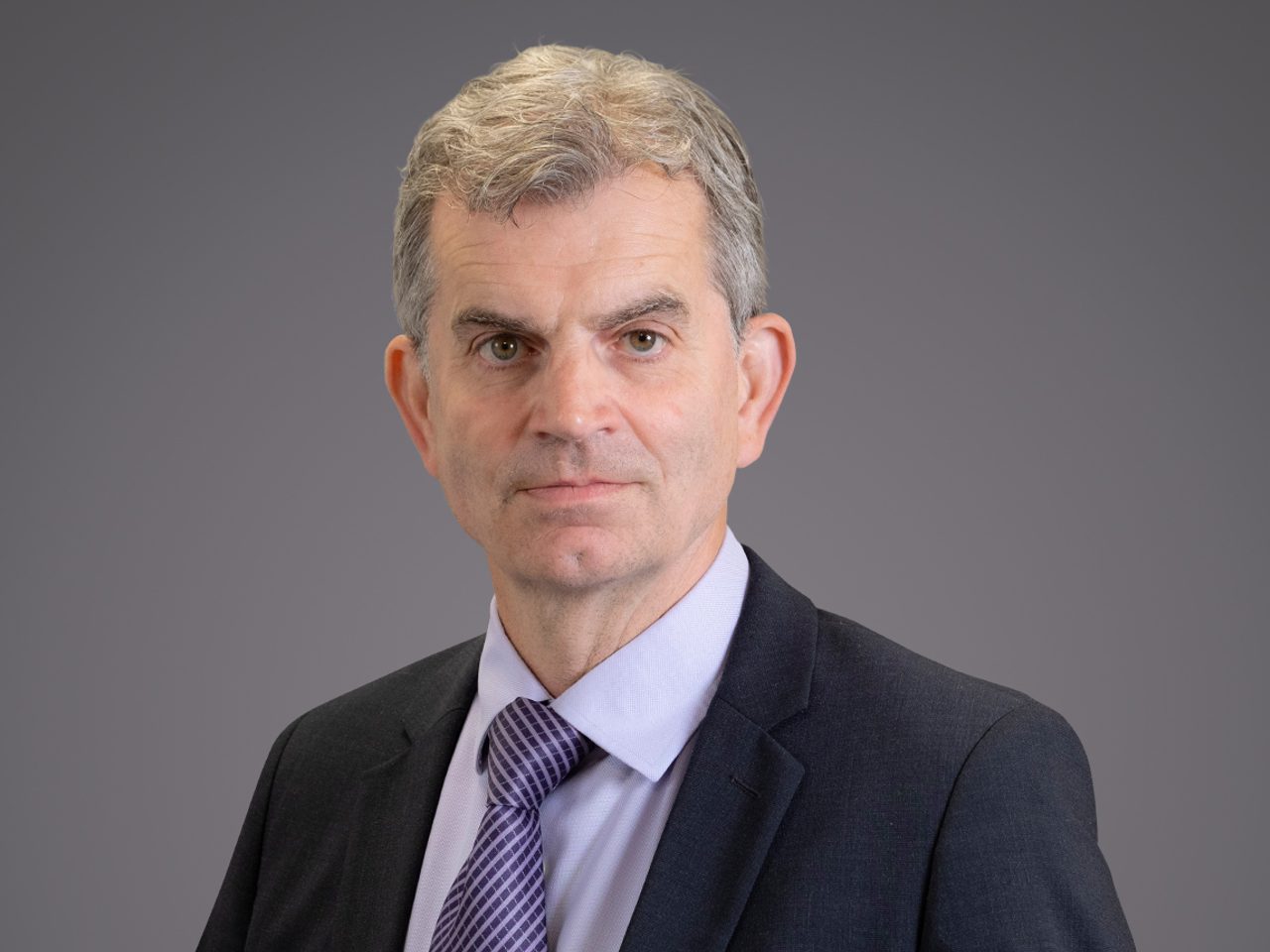 Portrait of a person with short grey hair, wearing a dark suit, light shirt and purple tie, facing the camera against a grey backdrop.