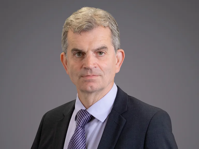Portrait of a person with short grey hair, wearing a dark suit, light shirt and purple tie, facing the camera against a grey backdrop.