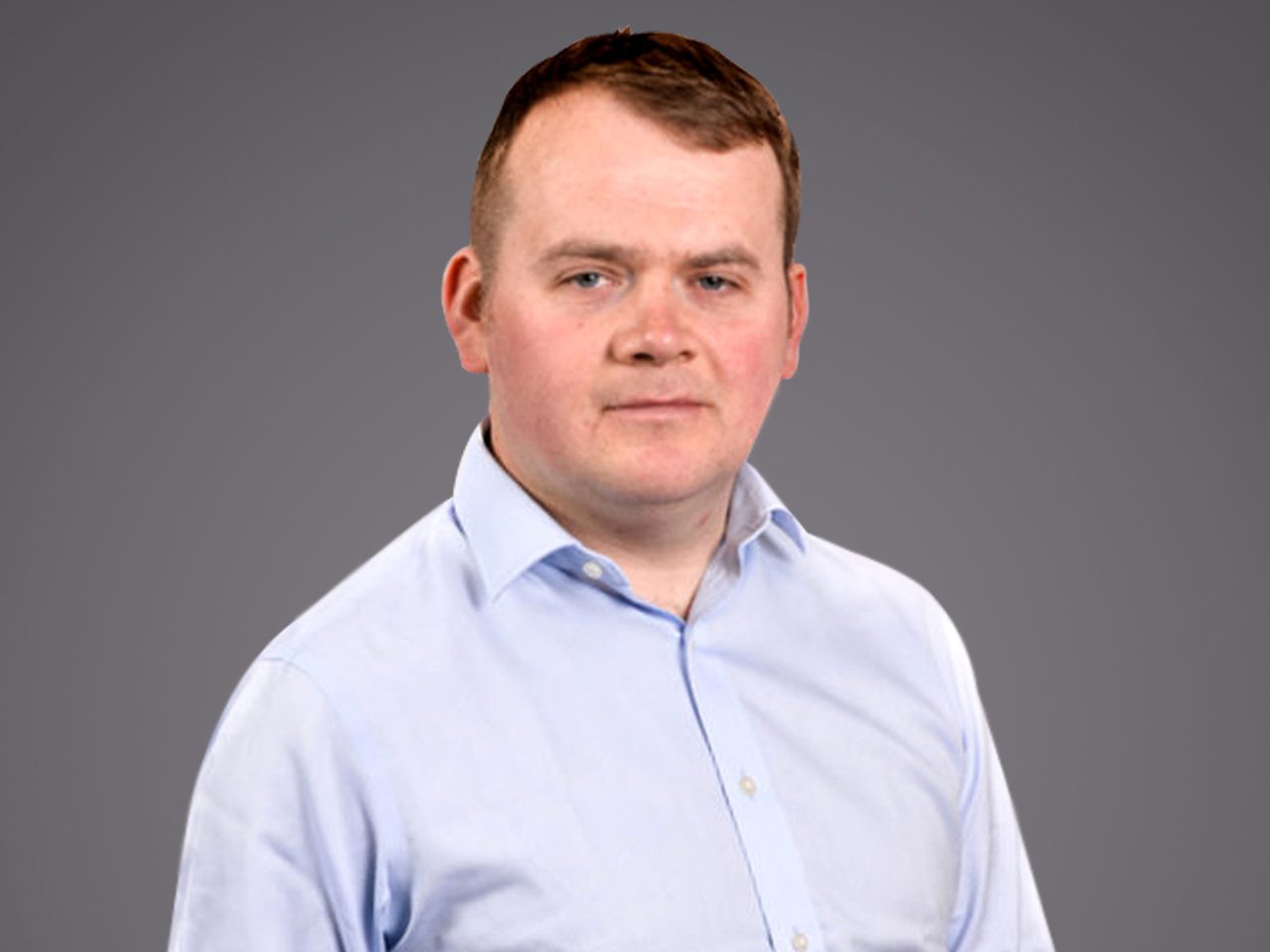 Portrait of a person with short brown hair wearing a light blue shirt, neutral expression, against a plain grey studio background.