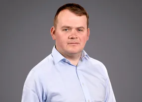 Portrait of a person with short brown hair wearing a light blue shirt, neutral expression, against a plain grey studio background.