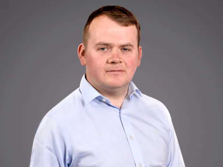 Portrait of a person with short brown hair wearing a light blue shirt, neutral expression, against a plain grey studio background.