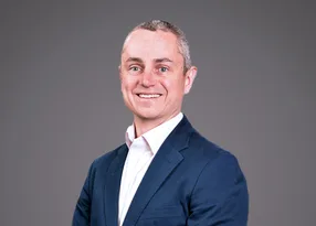 Smiling person in a navy blazer and white shirt with short grey hair, posed against a plain grey studio backdrop.