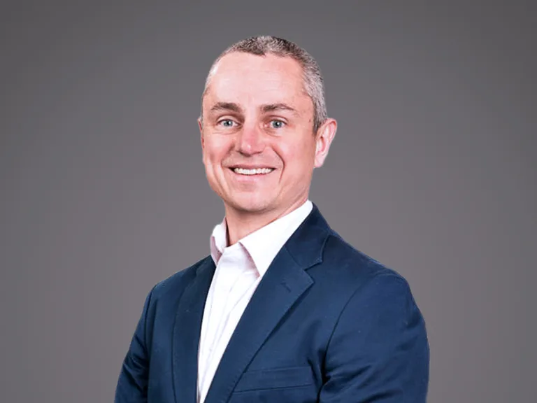 A smiling person wearing a navy blazer and white dress shirt, looking at the camera, against a solid gray studio backdrop.
