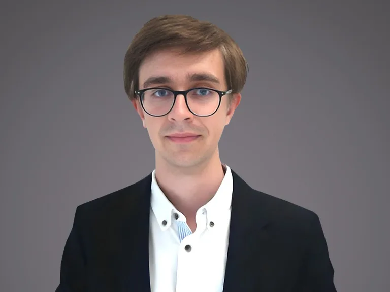 Portrait of a person with short brown hair and round glasses, wearing a white shirt and black blazer, against a neutral grey gradient background.