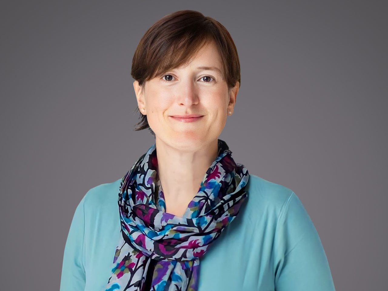 Portrait of a person with short brown hair, wearing a turquoise top and a multicoloured scarf, smiling softly against a grey backdrop.
