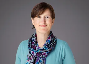 Portrait of a person with short brown hair, wearing a turquoise top and a multicoloured scarf, smiling softly against a grey backdrop.