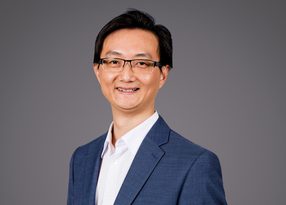 Portrait of a person with short black hair and glasses, wearing a blue blazer over a white shirt, smiling against a grey backdrop.
