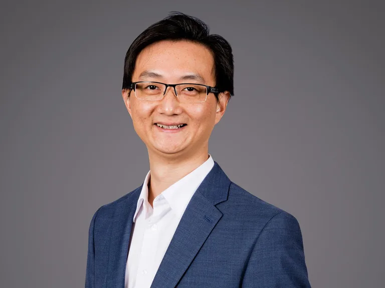 Portrait of a person with short black hair and glasses, wearing a blue blazer over a white shirt, smiling against a grey backdrop.