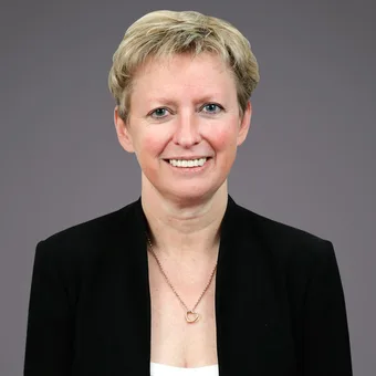Portrait of a person with short blond hair, wearing a black blazer and white top, smiling, with a heart pendant necklace, grey background.