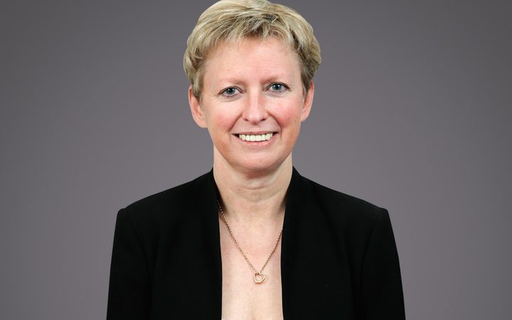 Portrait of a person with short blond hair, wearing a black blazer and white top, smiling, with a heart pendant necklace, grey background.