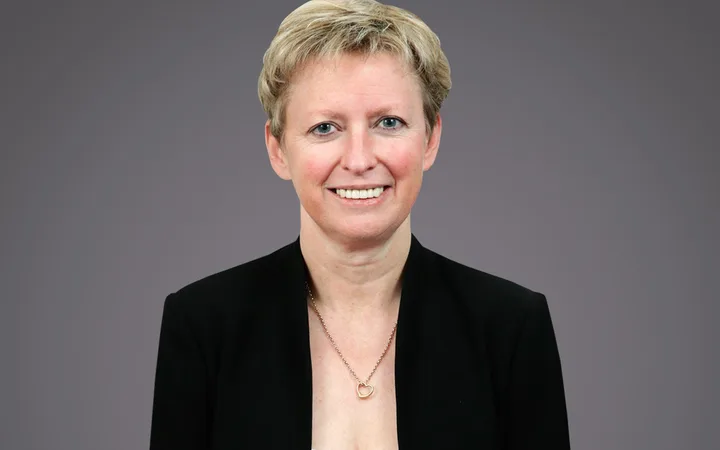 Portrait of a person with short blonde hair, wearing a black blazer and white top, smiling, with a heart pendant necklace against a gray backdrop.
