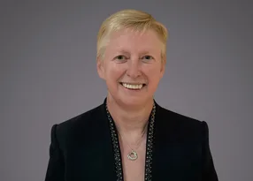 Smiling person with short blond hair wearing a black blazer with a beaded trim and a circular pendant necklace, against a grey background.