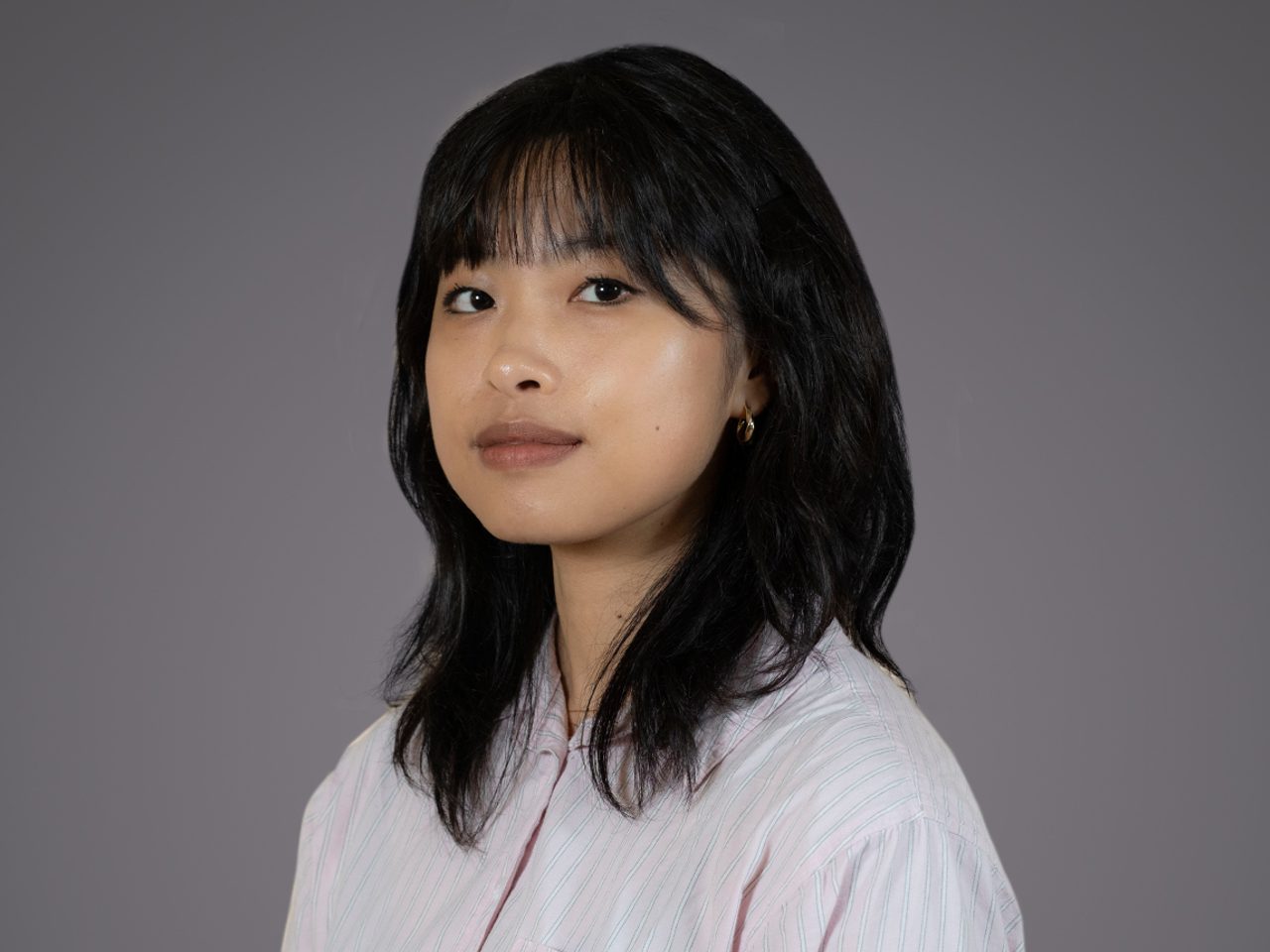 Portrait of an individual with shoulder-length black hair, wearing a pale pink striped shirt and small hoop earrings, neutral expression, gray background.