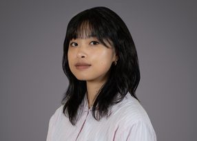 Portrait of an individual with shoulder-length black hair, wearing a pale pink striped shirt and small hoop earrings, neutral expression, gray background.