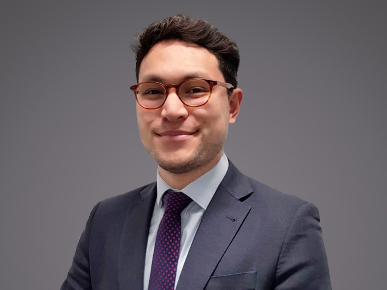 Portrait of a person in a dark suit, light shirt and purple dotted tie, wearing round glasses, smiling against a grey backdrop.
