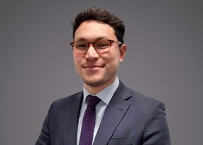 Portrait of a person in a dark suit, light shirt and purple dotted tie, wearing round glasses, smiling against a grey backdrop.