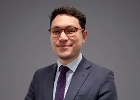 Portrait of a person in a dark suit, light shirt and purple dotted tie, wearing round glasses, smiling against a grey backdrop.