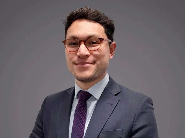 A person with short dark hair and round glasses, wearing a navy suit, light shirt, and purple dotted tie, smiling against a neutral gray background.