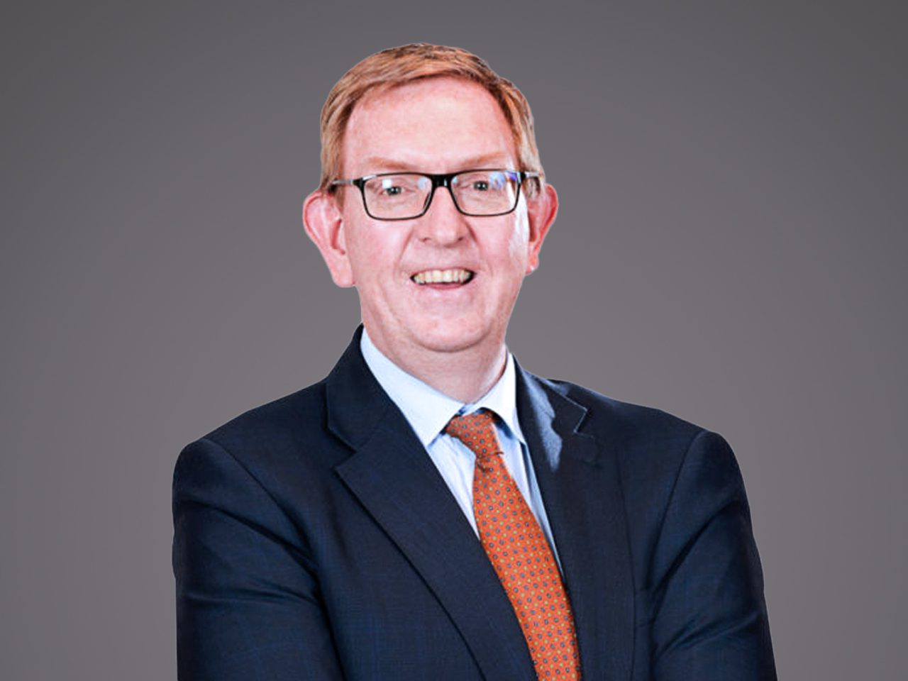 Person with short light hair and glasses, in a navy suit and orange patterned tie, smiling at the camera against a grey backdrop.