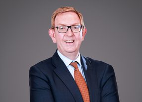 Person with short light hair and glasses, in a navy suit and orange patterned tie, smiling at the camera against a grey backdrop.
