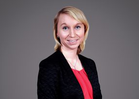 Portrait of a person with blonde hair, wearing a black patterned blazer and red blouse, smiling, against a grey backdrop.