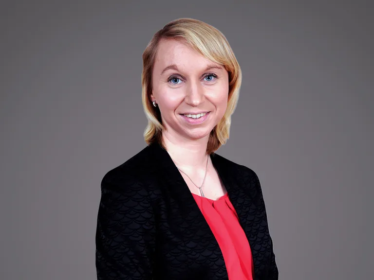 Portrait of a person with blonde hair, wearing a black patterned blazer and red blouse, smiling, against a grey backdrop.