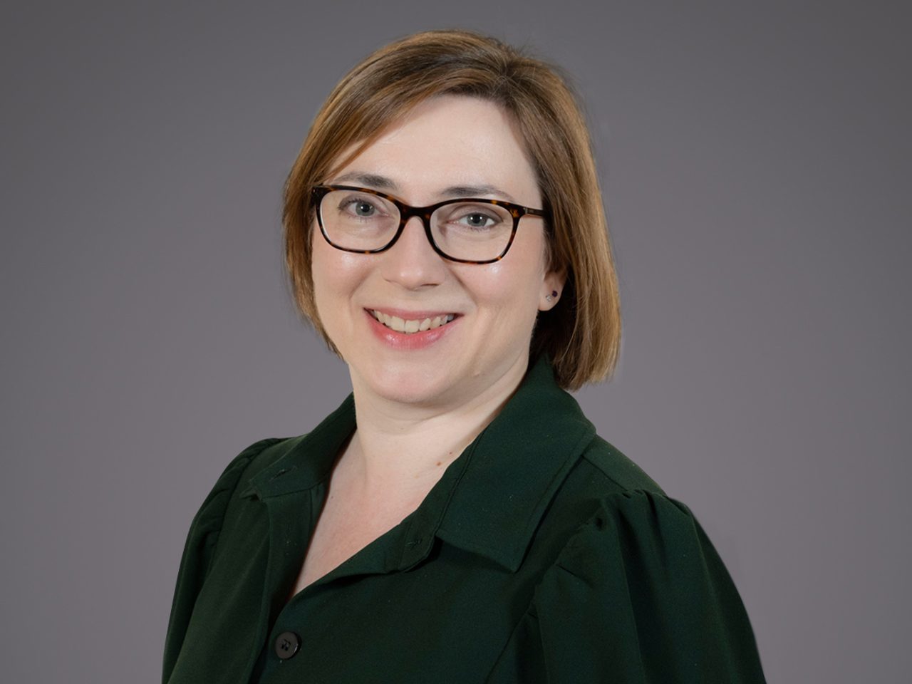 Person with short light brown hair and dark-framed glasses, wearing a dark green blazer, smiling against a grey studio backdrop.