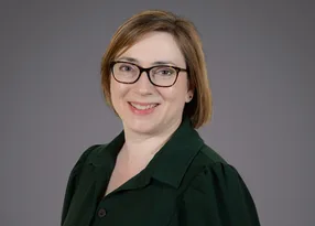 Person with short light brown hair and dark-framed glasses, wearing a dark green blazer, smiling against a grey studio backdrop.