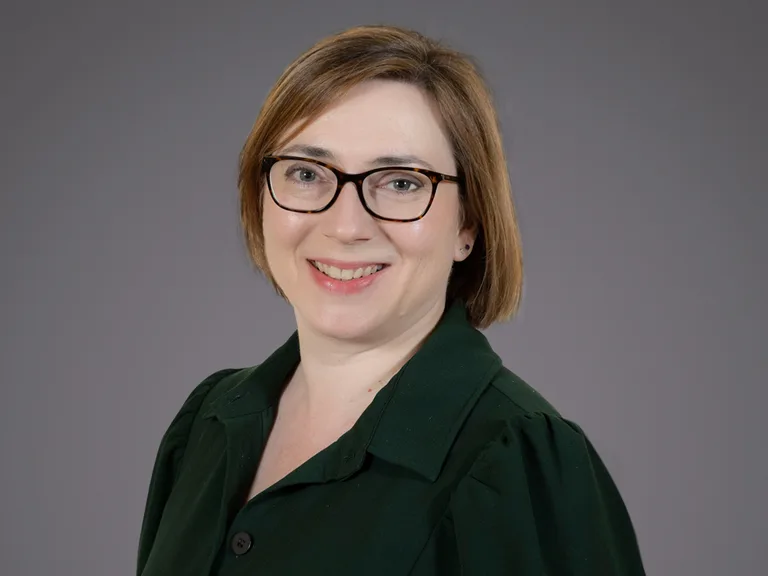 Person with short light brown hair and dark-framed glasses, wearing a dark green blazer, smiling against a grey studio backdrop.