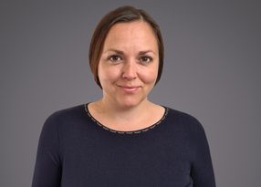 A person with short brown hair and a gentle smile, wearing a dark navy top with a stitched neckline, posed against a grey gradient background.