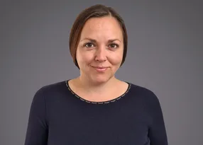 A person with short brown hair and a gentle smile, wearing a dark navy top with a stitched neckline, posed against a grey gradient background.