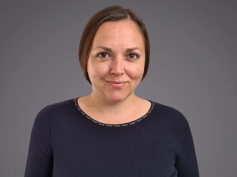 A person with short brown hair and a gentle smile, wearing a dark navy top with a stitched neckline, posed against a grey gradient background.