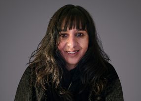 Portrait of a person with long dark hair and bangs, smiling, wearing a dark fur-trimmed coat, against a neutral grey background.