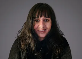 Portrait of a person with long dark hair and bangs, smiling, wearing a dark fur-trimmed coat, against a neutral grey background.