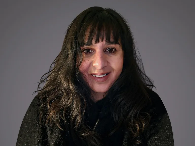 Portrait of a person with long dark hair and bangs, smiling, wearing a dark fur-trimmed coat, against a neutral grey background.