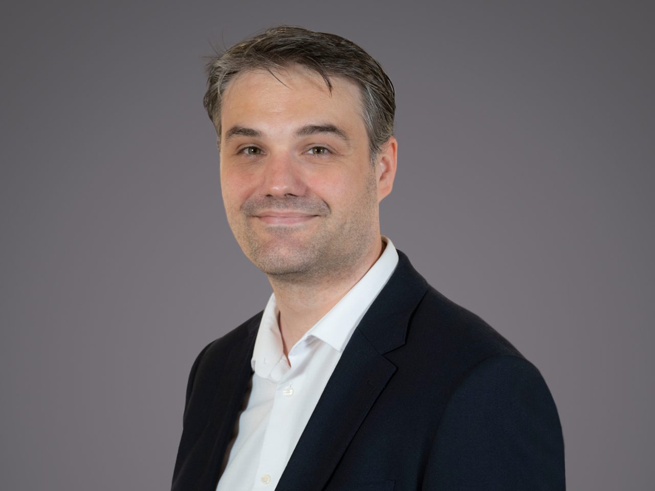 Portrait of a person with short dark hair, wearing a dark blazer and white shirt, against a grey backdrop, smiling softly.