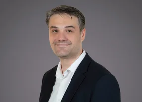 Portrait of a person with short dark hair, wearing a dark blazer and white shirt, against a grey backdrop, smiling softly.