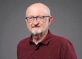 Portrait of an older person with short white hair and beard, wearing glasses and a maroon polo shirt, against a grey backdrop.