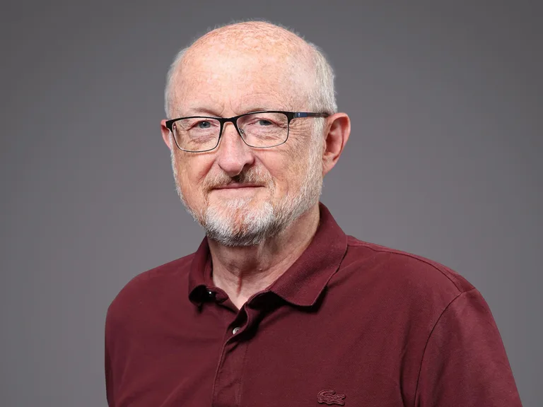 Portrait of an older person with short white hair and beard, wearing glasses and a maroon polo shirt, against a grey backdrop.