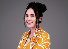 Person with dark hair in a messy bun, wearing an orange and white patterned shirt and a round pendant necklace, smiling at the camera against a grey backdrop.