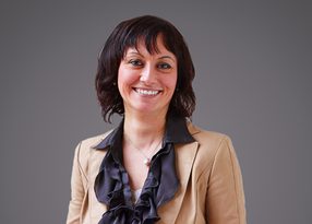 Smiling person with short dark hair, wearing a tan blazer and black ruffled blouse, against a grey backdrop.