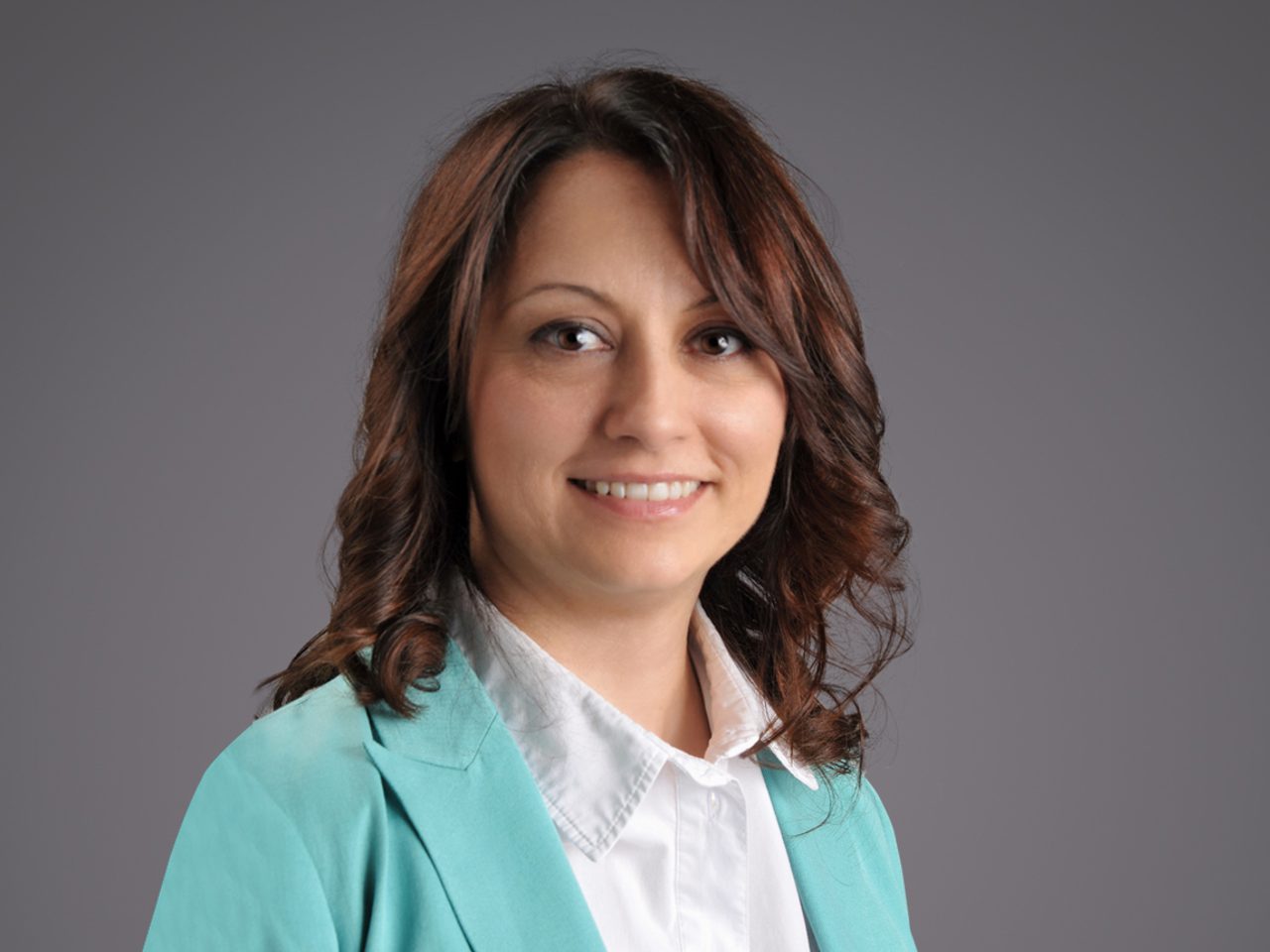 Head-and-shoulders portrait of a person with shoulder-length brown hair, wearing a white shirt and teal blazer, smiling against a gray backdrop.