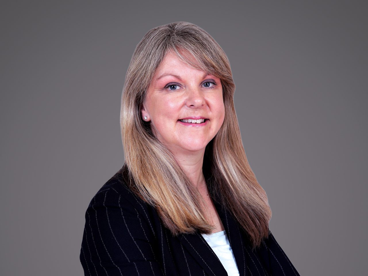 Professional portrait of a person with long blonde hair, wearing a pinstriped black blazer over a light top, smiling against a neutral grey backdrop.